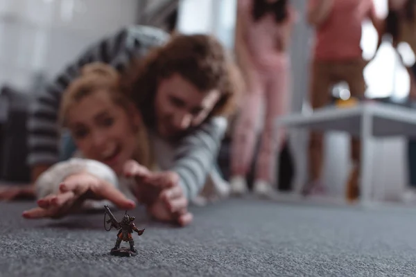 Selective focus of excited girl and guy lying on floor and trying to take toy figurine, and friends standing on background — Stock Photo