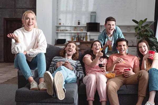 Amigos alegres sorrindo enquanto sentado no sofá em casa e assistindo tv — Fotografia de Stock