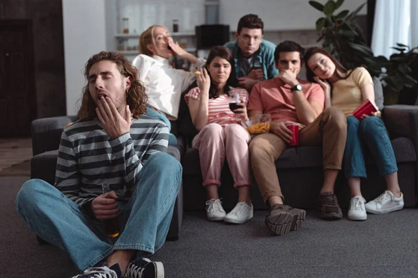 Selektiver Fokus junger Mann gähnt, während er auf dem Boden neben gelangweilten Freunden sitzt, die zu Hause fernsehen — Stockfoto