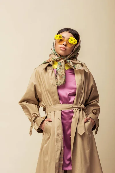 Trendy african american girl in sunglasses with flowers, head scarf and trench coat standing with hands in pockets isolated on beige — Stock Photo