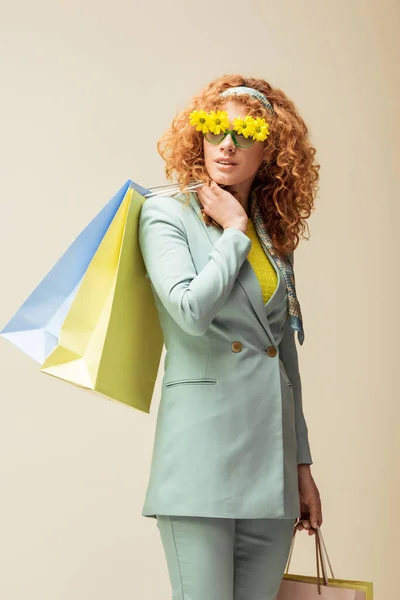 Elegante donna rossa in occhiali da sole con fiori che tengono borse della spesa isolate sul beige — Foto stock