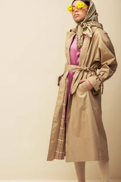 Stylish african american girl in sunglasses with flowers, head scarf and trench coat standing with hands in pockets on beige — Stock Photo