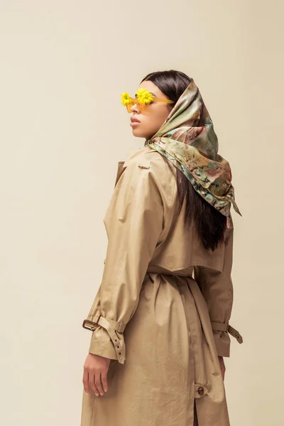 Стильная африканская американская девушка в солнцезащитных очках с цветами, шарфом на голове и пальто, изолированные на бежевом — стоковое фото