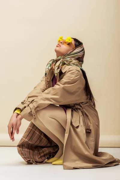 Stylish african american girl in sunglasses with flowers, head scarf and trench coat sitting on beige — Stock Photo