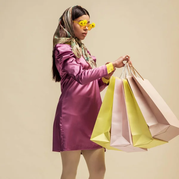 Trendy african american girl in sunglasses with flowers, head scarf and dress holding shopping bags isolated on beige — Stock Photo