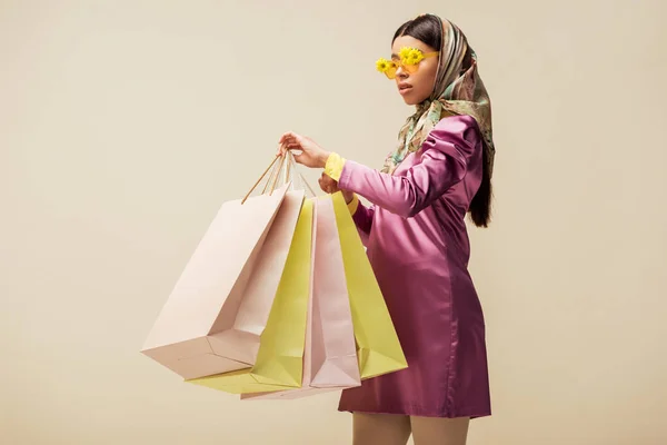 Stylish african american girl in sunglasses with flowers, head scarf and dress holding shopping bags isolated on beige — Stock Photo