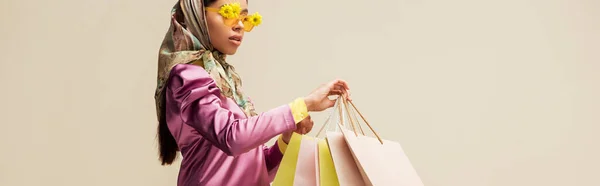 Tiro panorâmico da menina americana africana na moda em óculos de sol com flores e cachecol cabeça segurando sacos de compras isolados no bege — Fotografia de Stock