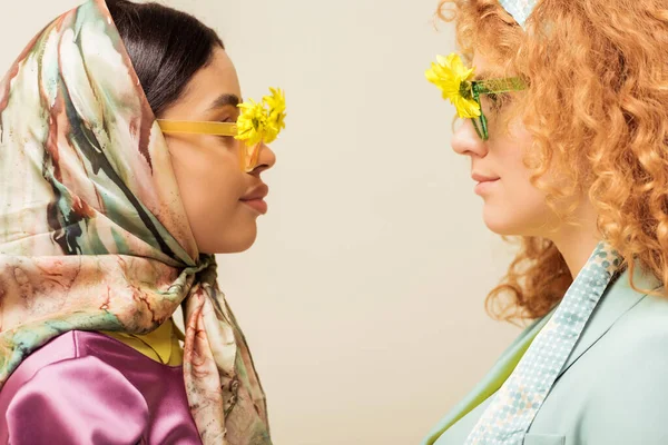 Vista lateral de chica afroamericana y pelirroja en gafas de sol con flores mirándose aisladas en beige - foto de stock