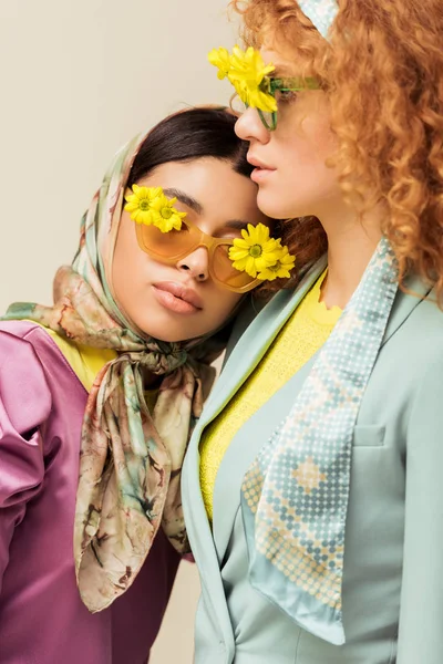 Chica afroamericana cerca de mujer pelirroja en gafas de sol con flores aisladas en beige - foto de stock