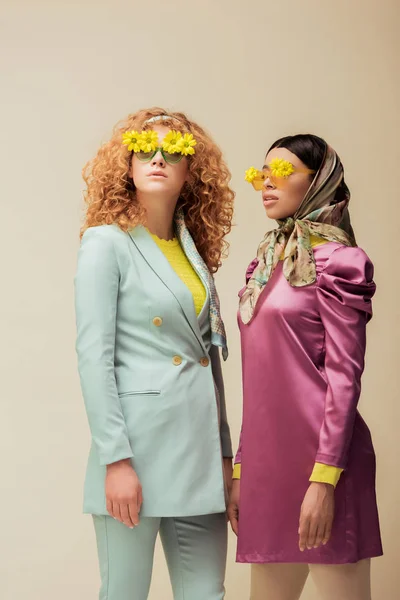 À la mode afro-américaine fille et rousse femme dans des lunettes de soleil avec des fleurs posant isolé sur beige — Photo de stock