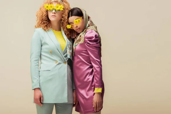 Chica afroamericana de moda y mujer pelirroja con estilo en gafas de sol con flores posando aisladas en beige - foto de stock