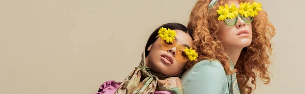 Tiro panorâmico da menina americana africana na moda e mulher ruiva em óculos de sol com flores posando isolado no bege — Fotografia de Stock