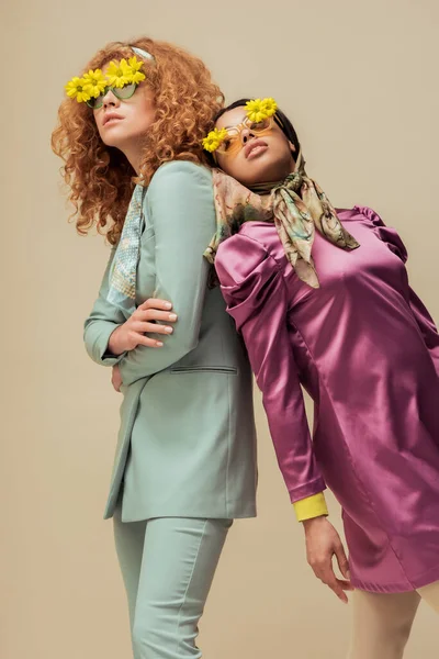 Ragazza afro-americana alla moda e donna riccia in occhiali da sole con fiori in posa isolata sul beige — Foto stock