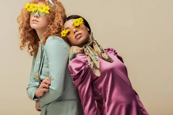 Élégante fille afro-américaine et femme bouclée dans des lunettes de soleil avec des fleurs posant isolé sur beige — Photo de stock