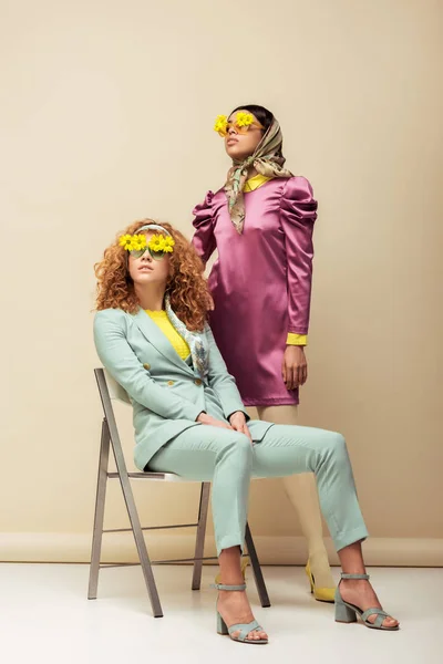 Mulher ruiva em óculos de sol com flores sentadas na cadeira perto de menina americana africana elegante posando no bege — Fotografia de Stock