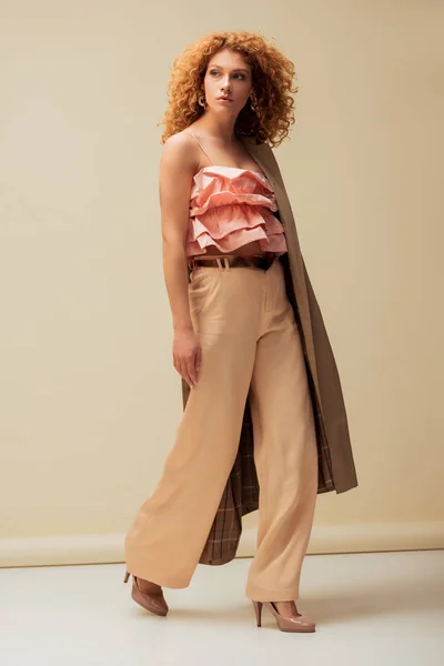Stylish redhead woman in ruffled top and trousers walking on beige — Stock Photo