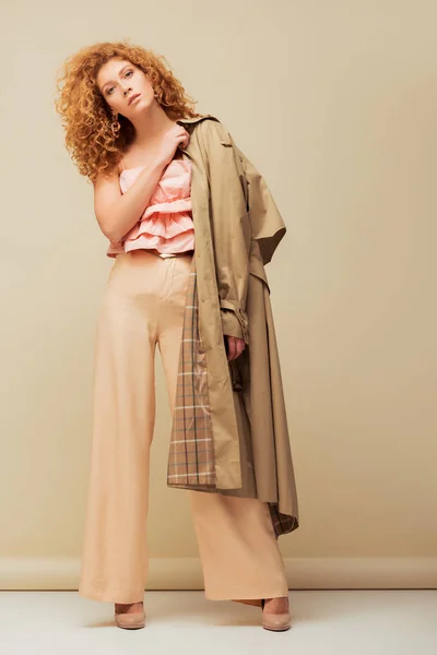 Stylish redhead woman in ruffled top and trousers holding trench coat while posing on beige — Stock Photo