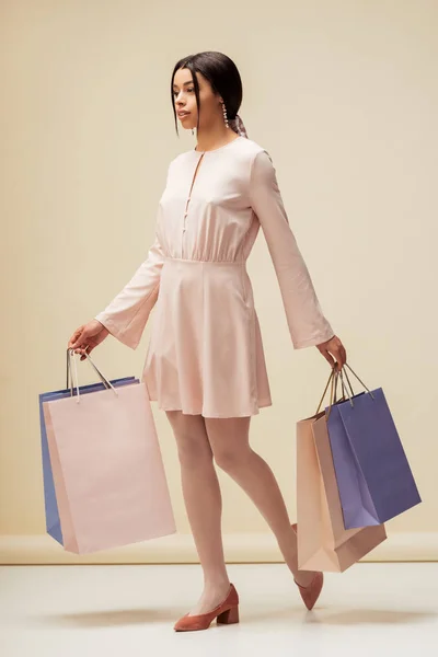 Attractive african american girl in dress holding shopping bags on beige — Stock Photo