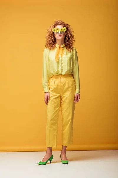 Кудрявая рыжая женщина в солнечных очках с цветами, позирующими на желтом — стоковое фото