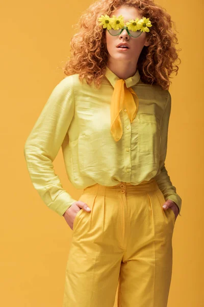 Curly redhead woman in sunglasses with flowers standing with hands in pockets isolated on yellow — Stock Photo