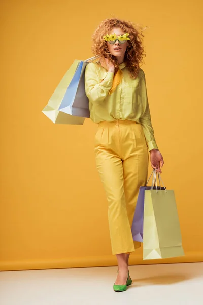 Stylische rothaarige Frau in Sonnenbrille mit Blumen, die Einkaufstüten halten und auf gelb posieren — Stockfoto