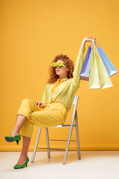 Stylische rothaarige Frau mit Sonnenbrille und Blumen, die Einkaufstüten hält, während sie auf gelb sitzt — Stockfoto