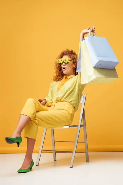 Excité rousse femme en lunettes de soleil avec des fleurs tenant des sacs à provisions sur jaune — Photo de stock