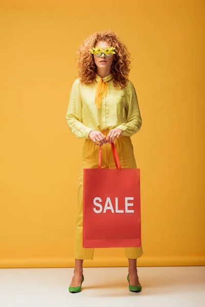 Elegante donna rossa in occhiali da sole con fiori in possesso di shopping bag con iscrizione in vendita su giallo — Foto stock
