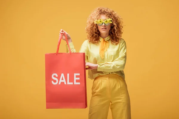 Élégant rousse femme en lunettes de soleil avec des fleurs tenant sac à provisions avec vente lettrage isolé sur jaune — Photo de stock