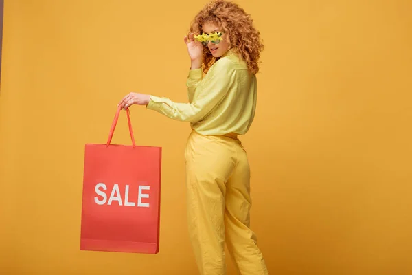 Élégant rousse femme touchant des lunettes de soleil avec des fleurs et tenant sac à provisions avec vente lettrage sur jaune — Photo de stock