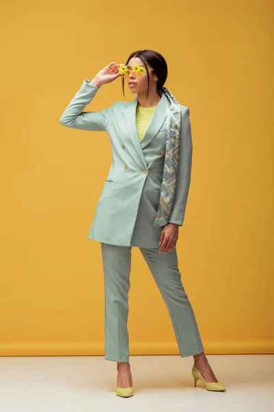 Trendy african american girl in suit and scarf touching sunglasses with flowers on yellow — Stock Photo