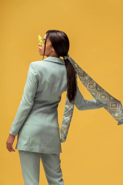 Vista laterale della ragazza afroamericana alla moda in giacca e cravatta, occhiali da sole con fiori e sciarpa isolata sul giallo — Foto stock