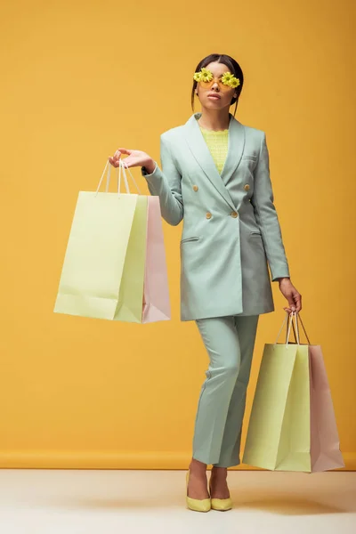 Atractiva chica afroamericana en traje y gafas de sol con flores sosteniendo bolsas de compras en amarillo - foto de stock