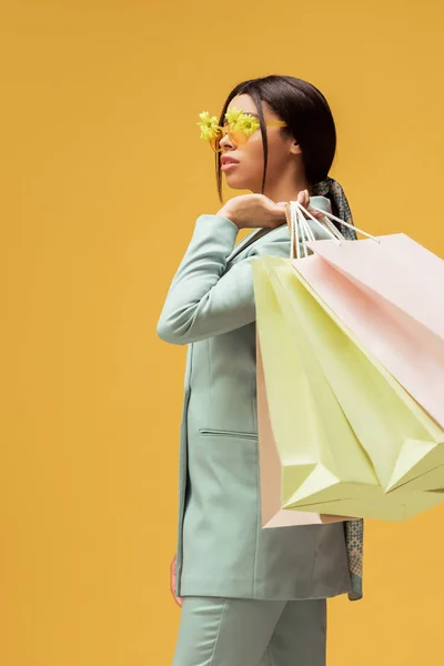 Attraktive afrikanisch-amerikanische Mädchen in Anzug und Sonnenbrille mit Blumen halten Einkaufstaschen isoliert auf gelb — Stockfoto