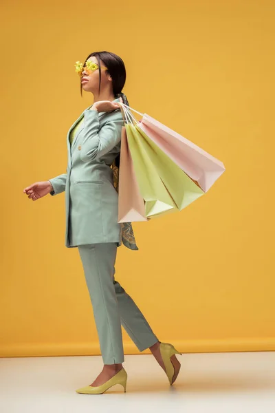 Atractiva mujer afroamericana en traje y gafas de sol con flores sosteniendo bolsas de compras en amarillo - foto de stock