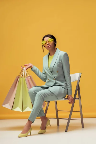 Atractiva mujer afroamericana en traje y gafas de sol con flores sosteniendo bolsas de compras mientras está sentado en amarillo - foto de stock