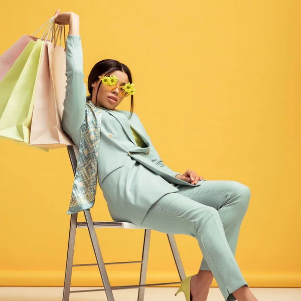 Stylish african american woman in suit and sunglasses with flowers holding shopping bags while sitting on yellow — Stock Photo