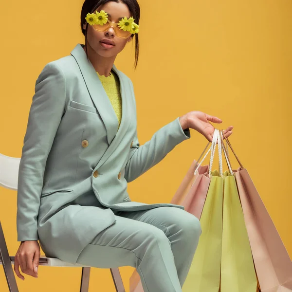 Elegante donna afroamericana in abito e occhiali da sole con fiori che tengono borse della spesa mentre seduto isolato sul giallo — Stock Photo