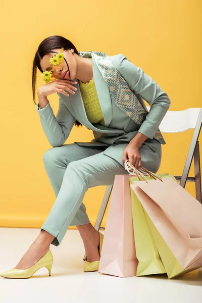 Trendy african american woman in suit, scarf and sunglasses with flowers holding shopping bags while posing on yellow — Stock Photo