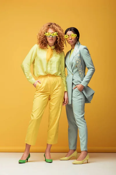 Atractivas mujeres multiculturales en gafas de sol con flores de pie con las manos en los bolsillos en amarillo - foto de stock