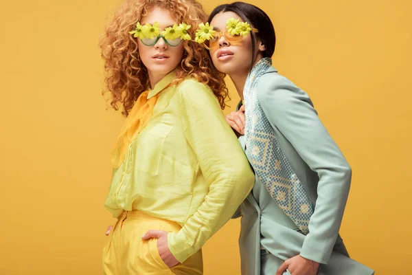Attractive multicultural women in sunglasses with flowers standing with hands in pockets isolated on yellow — Stock Photo
