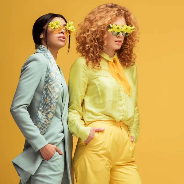 Довольно мультикультурных женщин в солнцезащитных очках с цветами стоя с руками в карманах изолированы на желтый — стоковое фото
