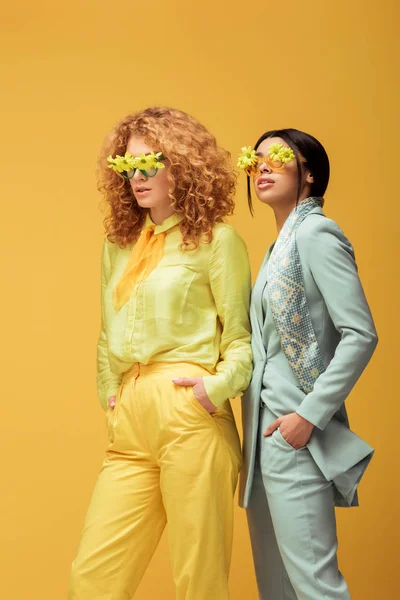 Belle donne multiculturali in occhiali da sole con fiori in piedi con le mani in tasche isolate sul giallo — Foto stock