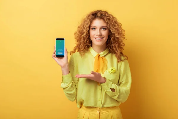 КИЕВ, УКРАИНА - 4 ФЕВРАЛЯ 2020: счастливая рыжая женщина, указывающая рукой на смартфон с приложением twitter на экране, изолированным на желтом — стоковое фото
