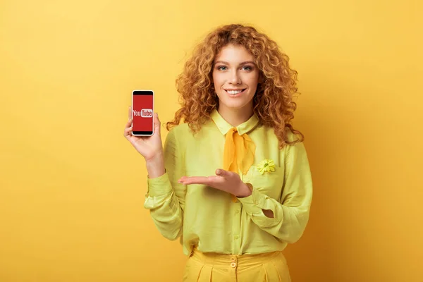 КИЕВ, УКРАИНА - 4 ФЕВРАЛЯ 2020: счастливая рыжая женщина, указывающая рукой на смартфон с приложением youtube на экране изолированы на желтый — стоковое фото