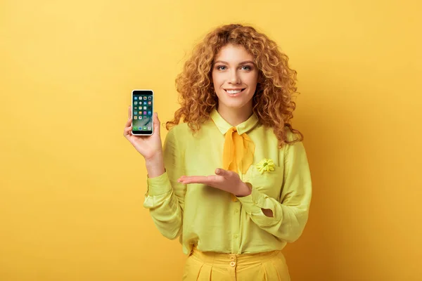 КИЕВ, УКРАИНА - 4 ФЕВРАЛЯ 2020: счастливая рыжая женщина, указывающая рукой на iphone изолирован на желтый — стоковое фото