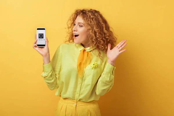 KYIV, UCRANIA - 4 de febrero de 2020: mujer pelirroja emocionada mirando el teléfono inteligente con la aplicación uber en amarillo - foto de stock