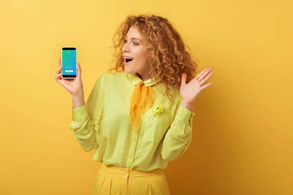 KYIV, UCRANIA - FEBRERO 4, 2020: mujer pelirroja emocionada mirando el teléfono inteligente con la aplicación de twitter en amarillo - foto de stock