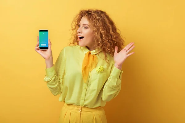 KYIV, UKRAINE - FEVEREIRO 4, 2020: mulher ruiva animado olhando para o smartphone com aplicativo twitter no amarelo — Stock Photo