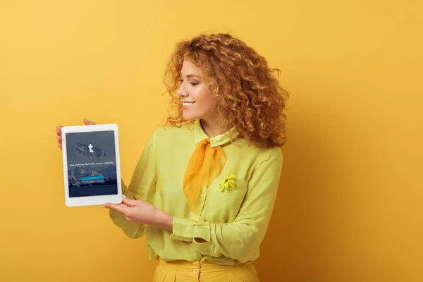 KYIV, UCRANIA - 4 de febrero de 2020: pelirroja alegre sosteniendo tableta digital con aplicación tumblr en amarillo - foto de stock